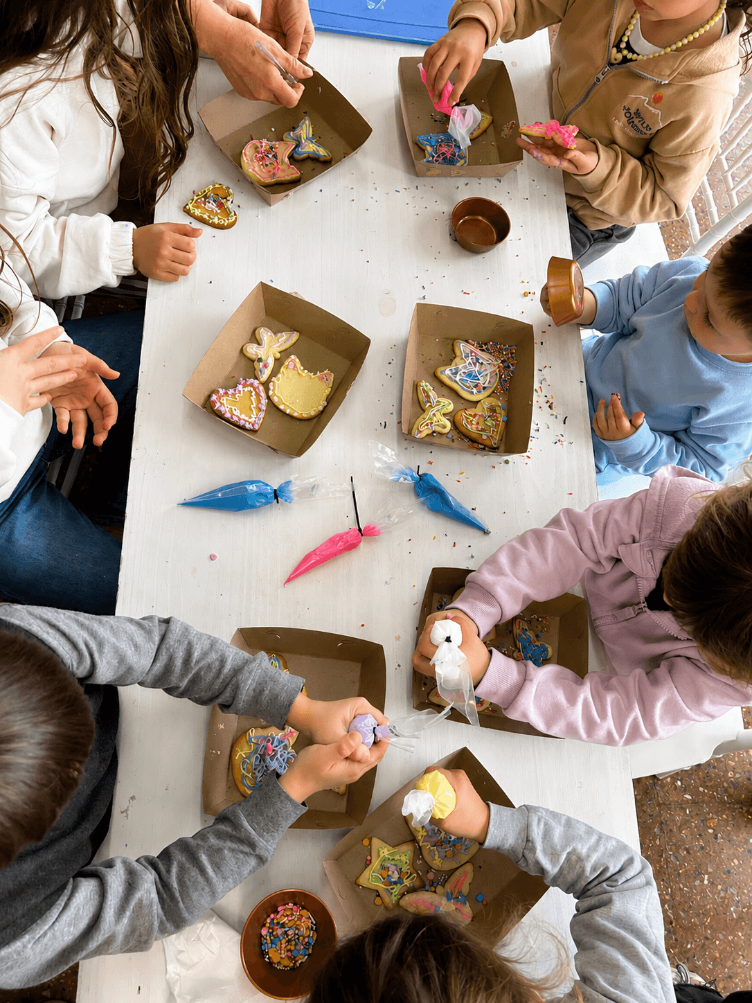 La mesa de una clase vista desde arriba: varias manos decorando galletitas, mangas de glasé y cajitas para llevar.