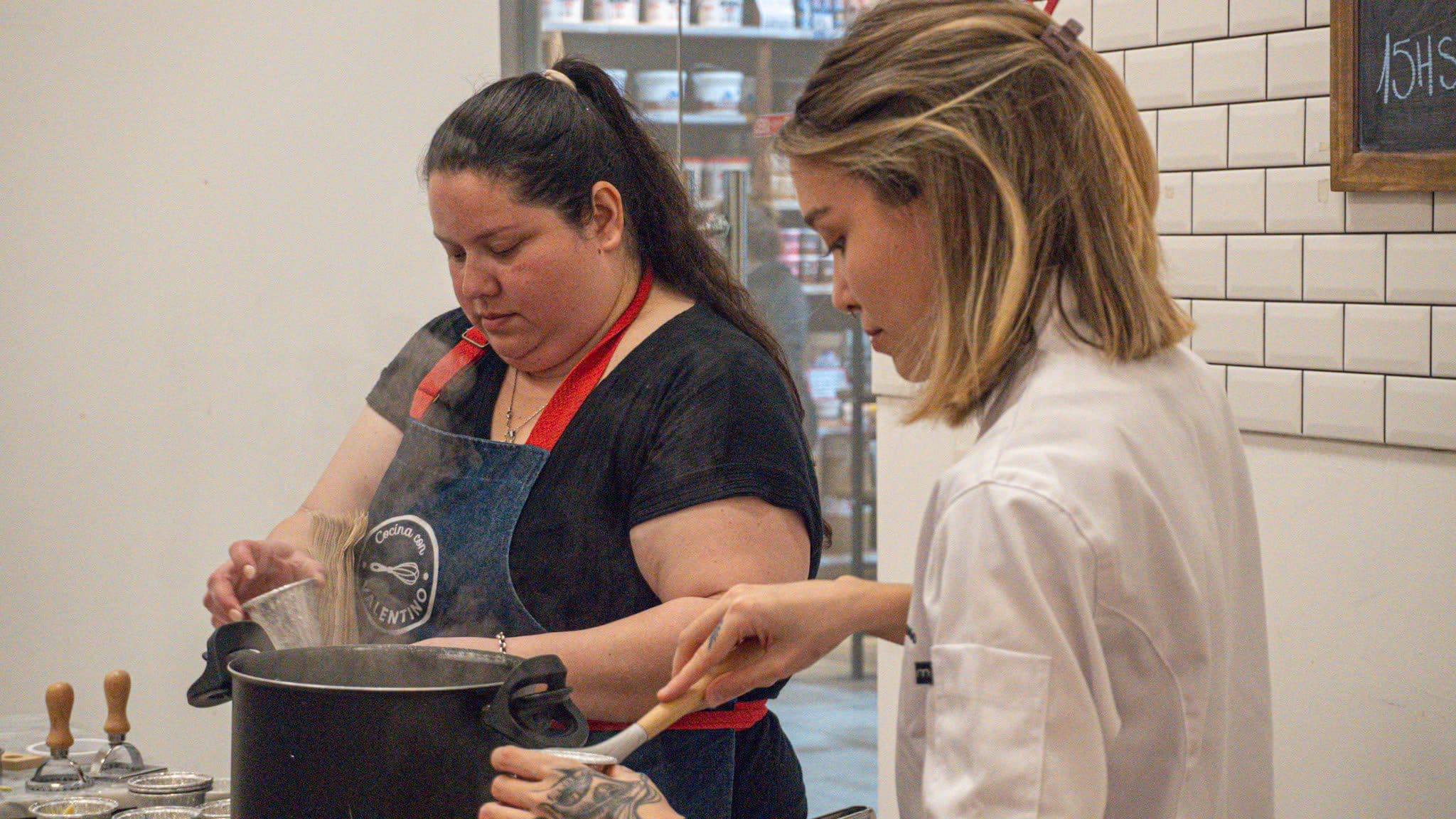 Delfina cocinando junto a otra persona: una revuelve la olla y la otra acompaña al lado.