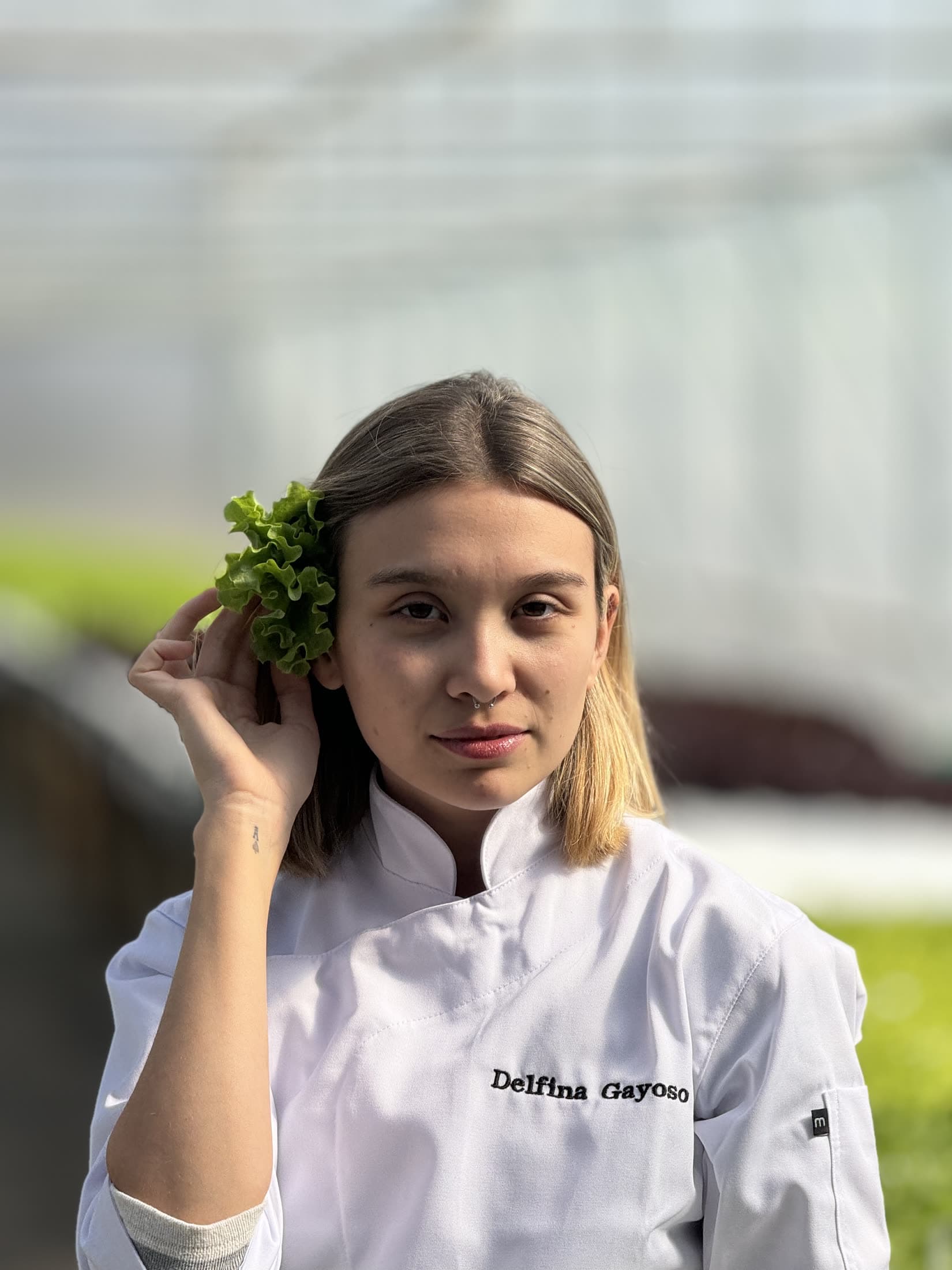 Delfina con la chaqueta de cocina, sosteniendo una hoja de lechuga junto a la sien, mirando a cámara.