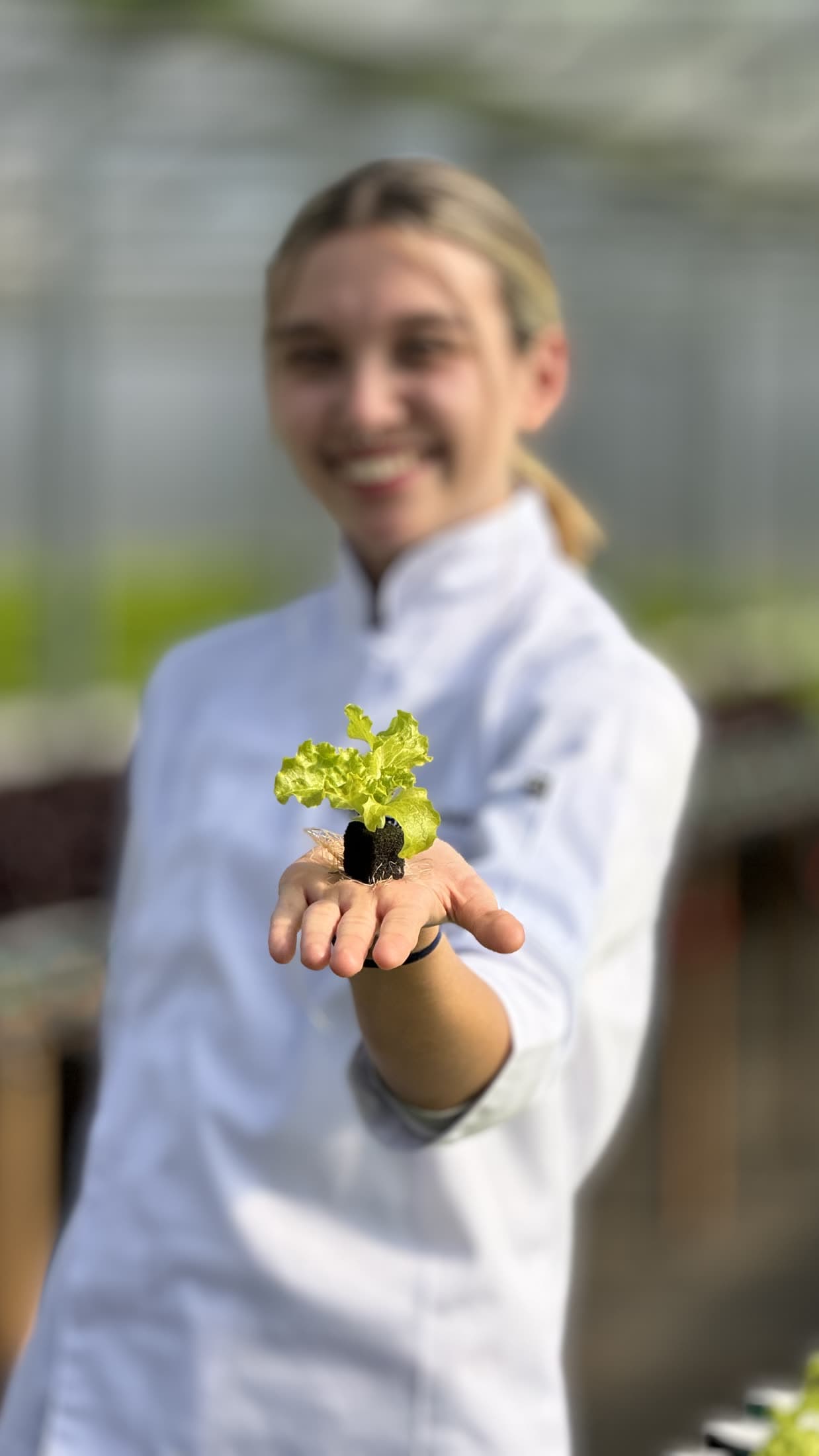 Delfina sonríe sosteniendo un brote de lechuga en la palma, al aire libre.
