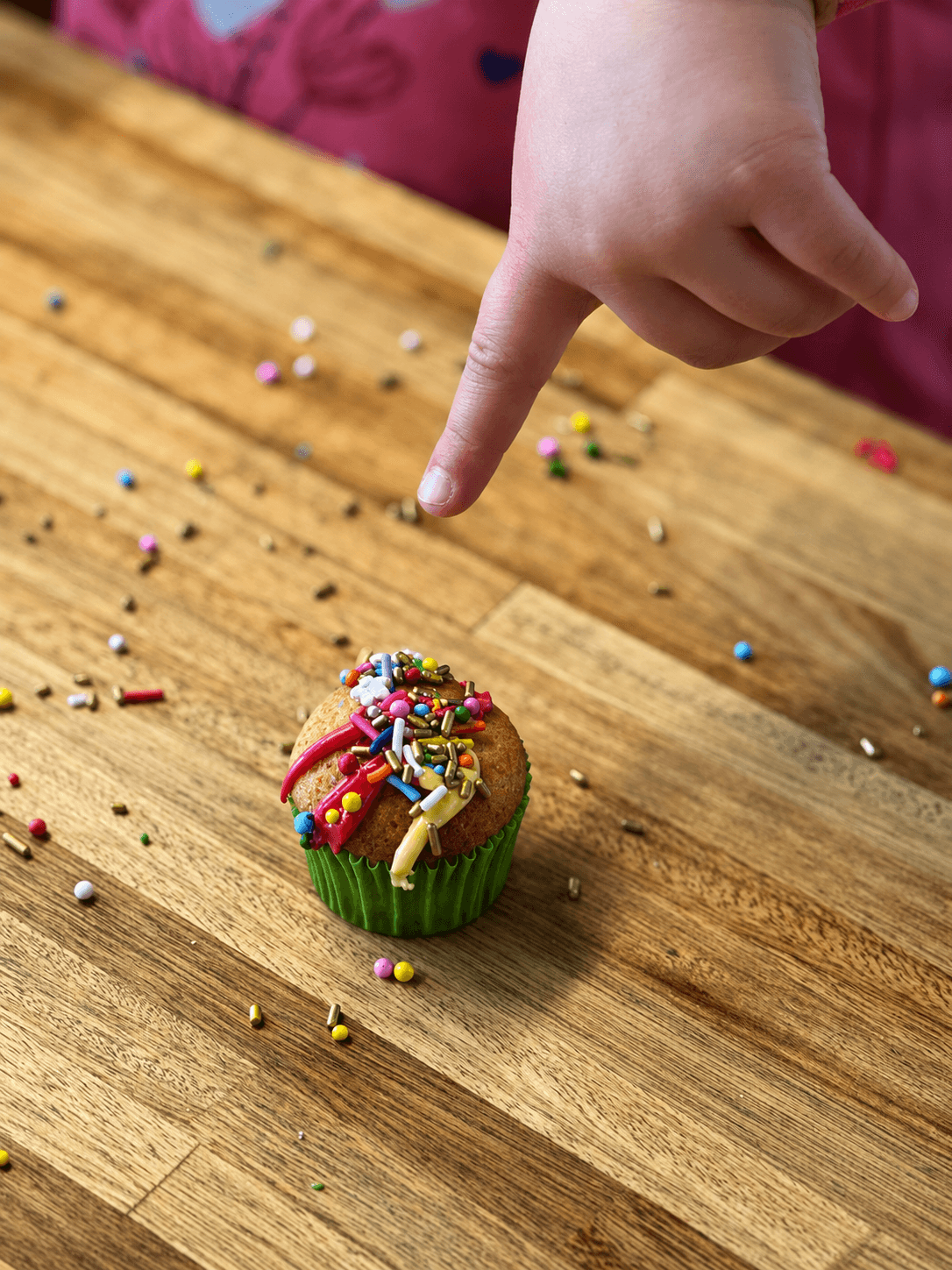 La mano de un chico señalando un cupcake decorado, con grana desparramada sobre la mesa de madera.