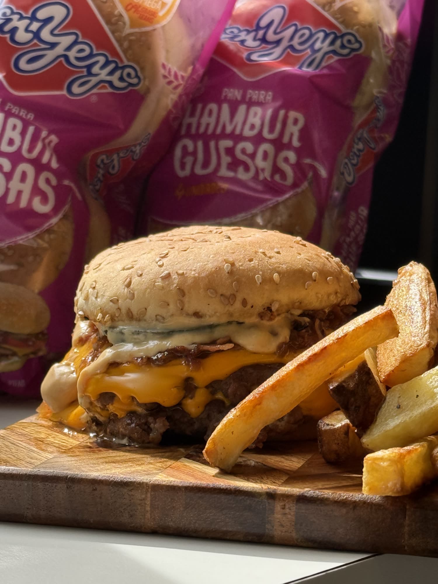 Una hamburguesa con papas rústicas sobre una tabla, con los paquetes de pan Don Yeyo detrás.