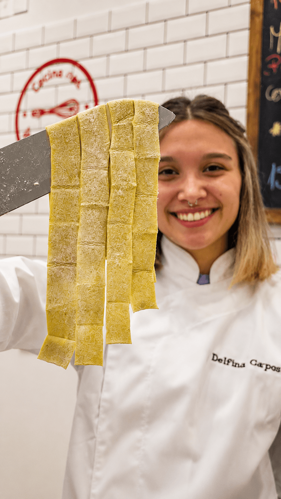 Delfina levanta con el cuchillo unas tiras de pasta verde recién cortadas, sonriendo detrás.