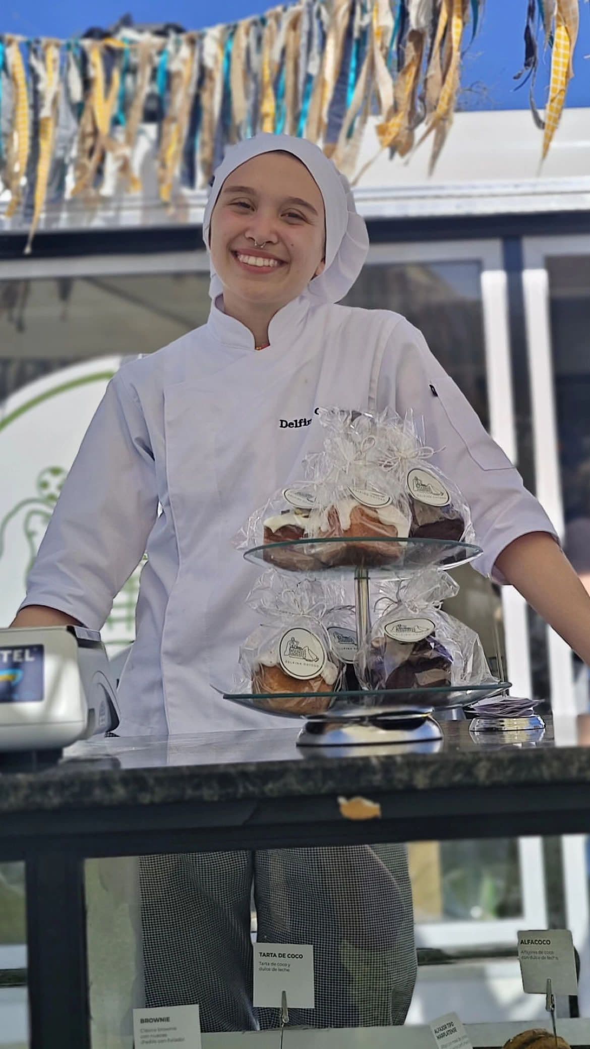 Delfina detrás del mostrador, con una bandeja de dos pisos y los adornos colgando.